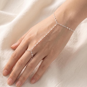 May include: A delicate silver hand chain featuring a fine, shimmering link design. The jewelry piece consists of a slender bracelet connected by a thin chain to a matching ring that encircles the middle finger, creating a continuous, elegant look.