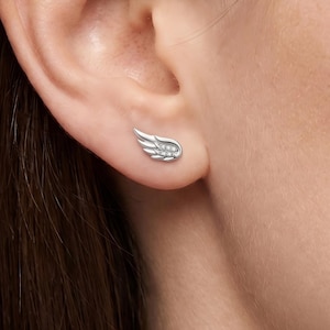 Può includere: Un orecchino a lobo color argento a forma di ala stilizzata. Il design dell'ala presenta segmenti metallici lisci e curvi ed è impreziosito da un piccolo gruppo di pietre chiare e scintillanti incastonate al centro della struttura.