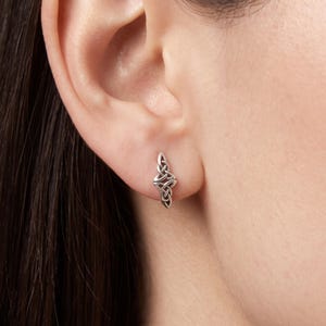 May include: A silver stud earring featuring an intricate, elongated Celtic triquetra knot design. The metal has a polished finish with darkened recessed areas that highlight the interwoven pattern of the trinity knot shape on the earlobe.