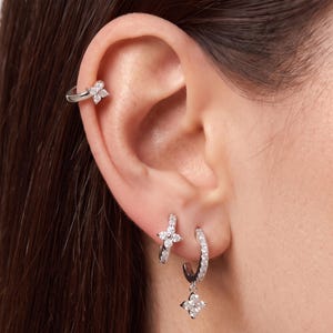 Puede incluir: Un ear cuff plateado con diseño de flor de cuatro pétalos y piedras transparentes, un pendiente de aro pequeño plateado con una flor a juego y un pendiente de aro plateado más grande con un dije colgante de flor de cuatro pétalos.