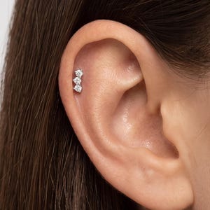 May include: A silver-toned cartilage earring featuring three small, round, clear stones arranged in a vertical row. The earring is secured in the upper ear, showcasing a polished metal setting that holds each stone in a prong-style mount.