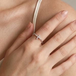 May include: A silver-toned ring featuring a delicate cross centrepiece adorned with small, clear, sparkling stones. The band is thin and metallic, designed to sit comfortably on the finger, showcasing a refined and polished appearance throughout.