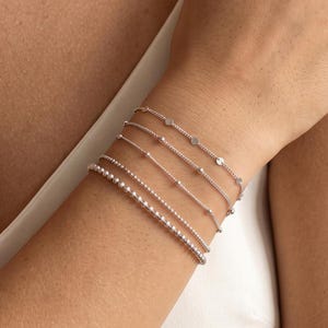 Peut inclure: Un ensemble de cinq bracelets argentés portés ensemble. La collection comprend une chaîne avec de petits disques circulaires plats, une chaîne délicate avec des perles espacées, une fine chaîne à maillons, une chaîne perlée et un bracelet en perles.