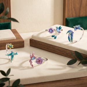 May include: Four silver-toned open rings featuring colorful enamel designs of hummingbirds and flowers. Each ring displays a unique combination of blue, pink, purple, and yellow floral motifs paired with small, detailed hummingbird figures.
