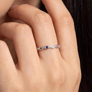 Op de afbeelding: Een zilverkleurige ring met een reeks markiesvormige metalen schakels, elk voorzien van een kleine ronde edelsteen. De stenen wisselen af in de kleuren groen, blauw, wit en roze, gezet in een gestructureerd, gepolijst metalen frame.