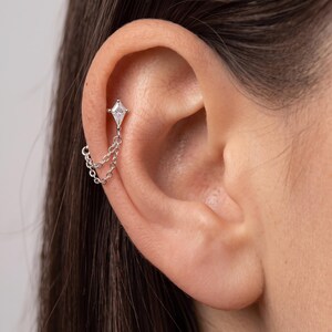 Puede incluir: Un pendiente de tipo ear cuff en tono plateado con un brillante transparente en forma de cometa, conectado a dos cadenas metálicas finas que cuelgan a lo largo del cartílago de la oreja. La pieza se sujeta al oído mediante un pequeño poste.