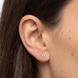May include: A small, silver-toned stud earring shaped like a V or chevron symbol. The earring features a polished, metallic finish and is positioned securely in the earlobe, showcasing a clean, geometric design that sits flat against the skin.