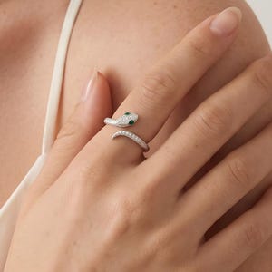 May include: A silver-toned wrap ring shaped like a snake, featuring a pavé-set surface with small clear stones and two small green stones for the eyes. The ring is designed as an open band that coils around the finger, showcasing a textured metallic finish.