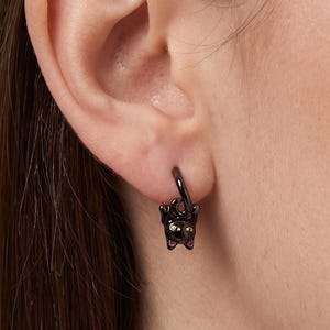 May include: A black hoop earring featuring a small, dangling black cat charm. The cat has gold-coloured eyes and small pink accents on the inner ears. The earring is constructed from a dark, polished metal, creating a sleek and uniform appearance.