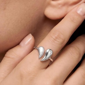 Puede incluir: Un anillo abierto de tono plateado con dos extremos en forma de corazón. Un extremo muestra tres pequeñas estrellas recortadas con piedras incrustadas, mientras que el otro presenta una luna creciente y la palabra Love grabada en cursiva.