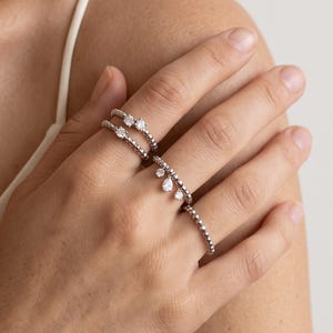 Peut inclure: Deux bagues perlées de couleur argentée portées aux doigts. La bague supérieure présente deux petites pierres rondes transparentes. La bague inférieure comporte trois pierres transparentes pendantes, dont une en forme de poire.