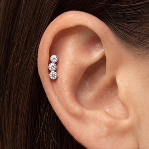 May include: A silver-toned ear cartilage piercing featuring three round, clear, bezel-set stones arranged in a vertical line. The jewelry is positioned along the upper edge of the ear, showcasing a clean, metallic finish and sparkling stones.