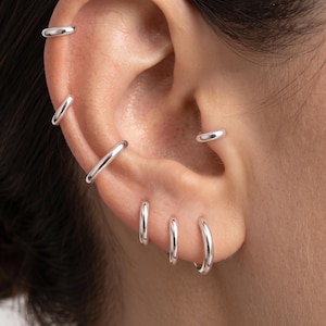 Op de afbeelding: Een set van zes gepolijste zilveren oorringen gedragen in verschillende oorpiercings, waaronder de helix, conch en oorlel. De ringen zijn glad, cirkelvormig en variëren licht in diameter, wat zorgt voor een gelaagde look op het kraakbeen.