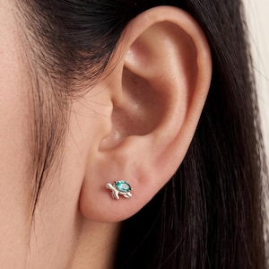 May include: A small silver-toned stud earring shaped like a turtle, featuring a single marquise-cut teal gemstone set into the shell. The earring is positioned on an earlobe, showcasing the metallic finish and the vibrant green-blue stone.