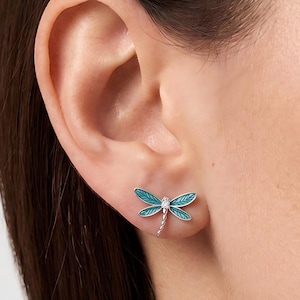 May include: A silver dragonfly stud earring featuring four wings filled with vibrant teal enamel and a small, clear, round-cut gemstone set at the centre of the body. The insect's tail is textured with a segmented design, and the wings have fine lines.
