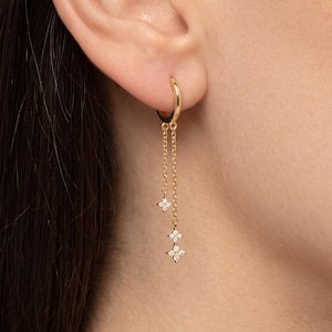 May include: A gold hoop earring featuring two delicate dangling chains of varying lengths. Each chain is accented with small, four-petalled flower-shaped charms encrusted with clear, sparkling stones. The earring is secured through the earlobe.