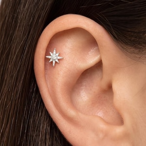 Puede incluir: Un pendiente de botón en forma de estrella de tono plateado con múltiples piedras pequeñas y claras dispuestas en un patrón radiante. La pieza de joyería está situada en la zona superior del cartílago de la oreja, con un diseño de varias puntas.