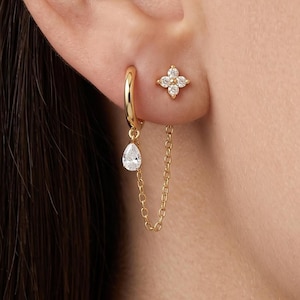 May include: A gold hoop earring with a dangling pear-shaped clear stone charm connected by a fine chain to a separate four-petaled flower stud earring featuring small clear stones. The jewellery set is crafted from polished gold-tone metal.