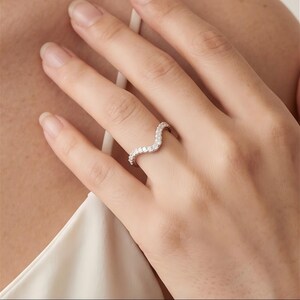 May include: A silver-toned chevron-shaped ring featuring a row of small, clear, round-cut stones set along the entire curved band. The ring has a polished metallic finish and follows a distinct wave-like contour that sits across the finger.