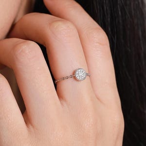 May include: A silver-coloured chain ring featuring a small, circular centrepiece paved with multiple tiny, clear, sparkling stones. The delicate band is constructed from fine metal links that wrap around the finger, creating a subtle and refined look.