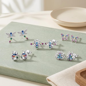 Pode incluir: Uma coleção de quatro pares de brincos de prata com designs de insetos e borboletas. Cada brinco é incrustado com pequenas pedras de zircônia cúbica multicoloridas em tons de azul, verde, amarelo, rosa e roxo, dispostas em padrões.