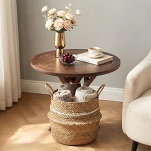 May include: A round wooden side table with a dark finish and a central pedestal base. On top sits a gold vase with light pink flowers, a small glass bowl of dark berries, and a white ceramic cup on a book. A woven seagrass basket holds folded blankets.