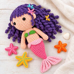 Könnte beinhalten: Eine gehäkelte Meerjungfrauenpuppe mit lockigem violettem Haar, das mit kleinen blauen, rosa und gelben Seesternen verziert ist. Die Puppe hat pfirsichfarbene Haut, ein grünes Oberteil und einen rosa Schwanz. Drei gehäkelte Seesterne in Rosa, Gelb und Orange liegen daneben.