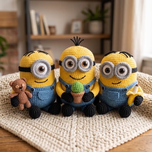 May include: Three crocheted yellow figures with black hair, gray goggles, and blue overalls. The left figure holds a small brown teddy bear, the center figure holds a green potted cactus, and the right figure holds a yellow banana. All have black feet.