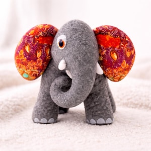 May include: A gray felt elephant plush toy featuring large, rounded ears with a vibrant red, orange, and purple patterned fabric. The elephant has a curled trunk, two small white tusks, and a single visible eye with a dark pupil and orange iris.
