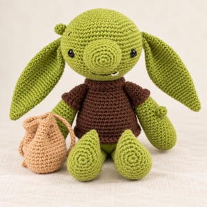 May include: A green crocheted creature doll with large ears, a round nose, and a single visible white tooth. The doll wears a brown short-sleeved shirt and is positioned next to a small, light brown crocheted drawstring pouch or bag.