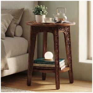 May include: A round wooden side table with intricate carved detailing on the legs and apron. The table features a lower shelf holding three stacked books titled Mediterranean Flora, Artisanal Textiles, and Historical Perspectives, topped by a small orb lamp.