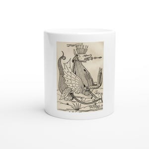 May include: A white ceramic mug featuring a black and white line art illustration of a mythical dragon-like creature. The creature has a crown-like headpiece, wings, and is depicted shooting a projectile from its mouth in a medieval woodcut style.