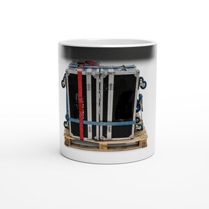 May include: A white ceramic mug featuring a graphic print of two black and silver road cases secured with blue and red straps on a wooden pallet. The cases have metal edges, corner protectors, and small wheels attached to the bottom of each unit.