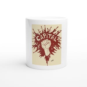 May include: A white ceramic mug featuring a graphic design on a cream-colored background. The design displays a dark red ink splatter shape with a raised fist in the center and the word CAPITAL printed in bold, distressed white capital letters.
