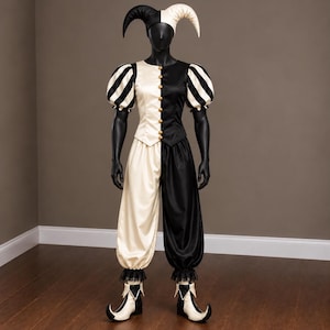 May include: A black and white jester costume featuring a two-toned bodice with gold buttons, puff sleeves, and matching loose-fitting trousers. The ensemble includes a two-pointed headpiece and pointed shoes, all designed in a split color pattern.