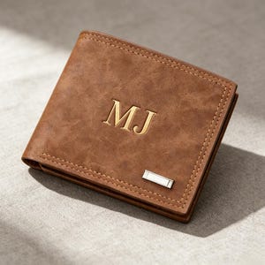 May include: A brown bi-fold wallet with a textured finish featuring the embossed letters MJ in the center. The wallet includes a small rectangular silver-toned metal accent piece on the lower right corner and visible stitching along the edges.