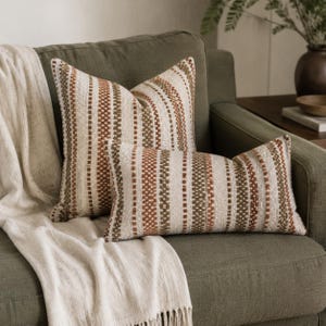 May include: Two decorative throw pillows featuring a textured, woven fabric with vertical stripes in shades of cream, rust orange, and olive green. One pillow is square, and the other is rectangular, both displaying a consistent pattern of alternating colors.