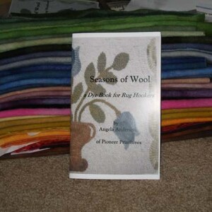 May include: A book titled "Seasons of Wool" is displayed in front of a stack of colourful wool fabric. The book cover features a floral design and the text "a Dye Book for Rug Hookers". The wool fabric is in shades of green, blue, pink, and orange.