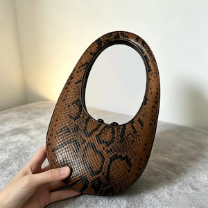 May include: A brown and black snakeskin patterned handbag with a distinct oval shape and a large integrated circular handle. The bag features a textured surface with a repeating reptilian scale print and a smooth dark interior lining visible.