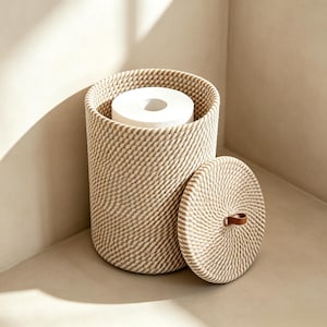 Puede incluir: Una cesta de almacenamiento cilíndrica hecha de cuerda trenzada en color beige y blanco, que contiene un rollo de papel higiénico blanco. La cesta incluye una tapa circular a juego con una pequeña asa de cuero marrón en el centro.