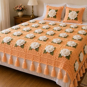 May include: A peach-colored crochet bedspread featuring a grid of white floral motifs with green leaves, finished with matching peach tassels along the edges. Two matching square crochet pillows with central white flowers sit on the bed.