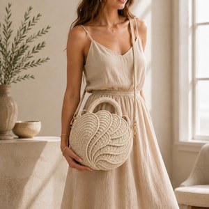 Peut inclure: Un sac à main rond en paille beige avec un motif texturé en forme de pétales ou d'ailes superposées sur le devant. Le sac est doté d'une poignée supérieure rigide et d'une longue bandoulière fine fixée par des attaches en métal doré.