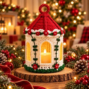 May include: A crocheted lantern cover featuring a red conical roof with a top loop, a cream-colored body with arched openings, and green trim. The design is decorated with small red flowers and green vine motifs, housing a single white pillar candle.