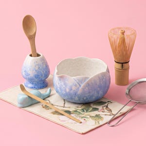 May include: A matcha tea set including a blue and white ceramic bowl with a scalloped rim, a matching whisk holder with a wooden spoon, a bamboo whisk, a bamboo scoop on a blue ceramic rest, and a stainless steel mesh strainer on a floral mat.