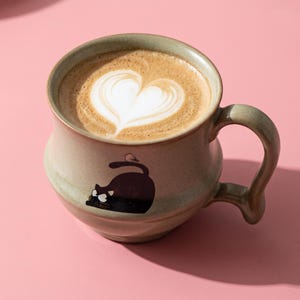 Puede incluir: Una taza de cerámica de color verde claro con forma redondeada y asa curva, llena de café con un diseño de arte latte en forma de corazón. En la parte frontal de la taza hay una ilustración de un gato negro con un pequeño pájaro en la cola.