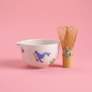May include: A white ceramic matcha bowl featuring a raised blue whale, a pink starfish, and green seaweed motifs, paired with a light brown bamboo matcha whisk adorned with a small blue seashell charm and a white faux pearl accent.