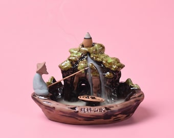 May include: A ceramic incense burner shaped like a mountain landscape with a small figure wearing a conical hat holding a fishing rod. The piece features green and brown glazes, a smoking incense cone at the peak, and Chinese characters.
