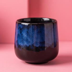 May include: A ceramic cup featuring a rounded silhouette with a glossy finish. The upper portion displays a vibrant, mottled blue glaze that transitions into a solid, deep black glaze covering the lower half and the base of the vessel.