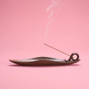 May include: A dark brown, elongated, curved incense holder with a smooth finish. A single incense stick is inserted into the circular holder at one end, with a thin trail of white smoke rising vertically into the air above the tray.