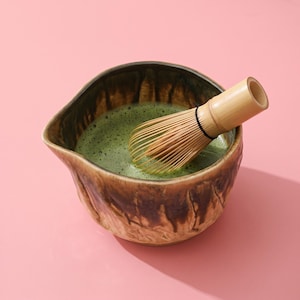 May include: A ceramic matcha bowl with a brown and tan drip glaze finish containing a vibrant green frothy tea. A light-colored bamboo whisk with thin tines rests inside the bowl, partially submerged in the liquid, angled toward the right side.
